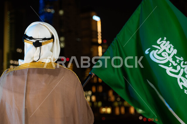 Founding of the Saudi state 1727 AD, Flag Day, March 11, celebration of the Saudi National Day, September 23, a close-up photo from the back of a Saudi Gulf Arab man wearing the bisht and ghutra on National Day, holding the Kingdom’s flag in his hand, standing in front of the illuminated towers and skyscrapers at night in Riyadh