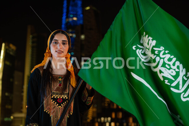 الاحتفال بذكرى اليوم الوطني السعودي 23 سبتمبر، أعياد ومناسبات وطنية ، يوم العلم 11 مارس، صورة مقربة لامرأة عربية خليجية سعودية ترتدي الثوب والشيلة الجنوبية في العيد الوطني تحمل بيدها علم المملكة، خلفية الابراج وناطحات السحاب المضيئة ليلا في الرياض