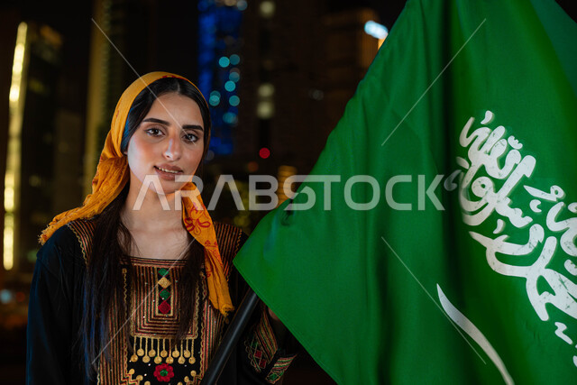 يوم العلم 11 مارس ،الاحتفال بذكرى اليوم الوطني السعودي 23 سبتمبر، احتفالات ومناسبات وطنية ، صورة مقربة لامرأة عربية خليجية سعودية مبتسمة ترتدي الثوب والشيلة الجنوبية في العيد الوطني تحمل بيدها علم المملكة، خلفية الابراج وناطحات السحاب المضيئة ليلا