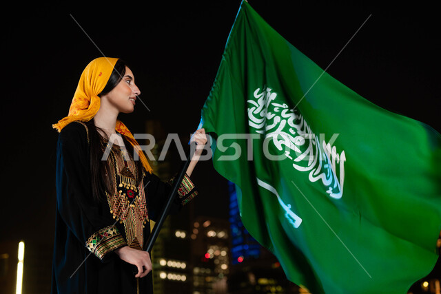 Highlighting love and loyalty to the country, national celebrations and events, commemorating the Saudi National Day, September 23, Flag Day, March 11, a Saudi Gulf Arab woman wearing the thobe and the southern shayla on National Day, holding the Kingdom’s flag in her hand with proud gestures, the background of skyscrapers illuminated at night in Saudi Arabia
