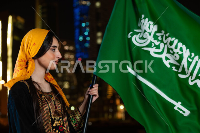 مشاعر الحب والانتماء للبلاد، احتفالات ومناسبات وطنية ، ذكرى اليوم الوطني السعودي 23 سبتمبر، يوم العلم 11 مارس، صورة مقربة لامرأة عربية خليجية سعودية ترتدي الثوب والشيلة الجنوبية في العيد الوطني تحمل بيدها علم المملكة، خلفية ناطحات السحاب المضاءة ليلا في السعودية