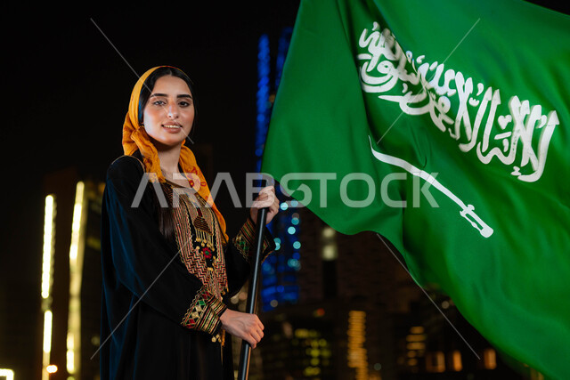 مشاعر الانتماء والحب للبلاد ،ذكرى اليوم الوطني السعودي 23 سبتمبر، احتفالات ومناسبات وطنية، يوم العلم 11 مارس، شابه عربية خليجية سعودية مبتسمة ترتدي الثوب والشيلة الجنوبية في العيد الوطني و تحمل بيدها علم المملكة، خلفية ناطحات السحاب المضيئة ليلا في الرياض