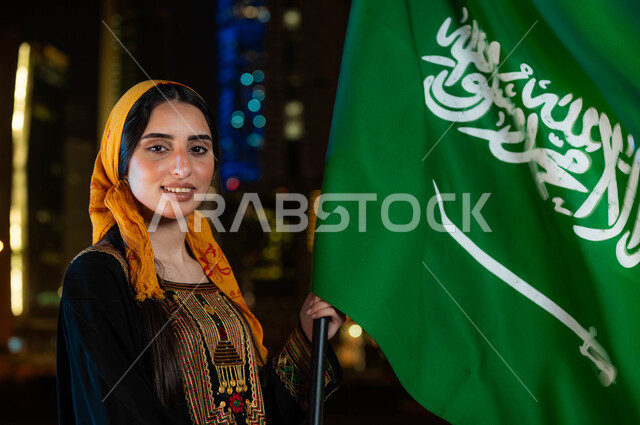 اظهار التفاني للبلاد ،الاحتفال بذكرى اليوم الوطني السعودي 23 سبتمبر، مناسبات و أعياد وطنية، يوم العلم 11 مارس، صورة مقربة لامرأة عربية خليجية سعودية مبتسمة ترتدي الثوب والشيلة الجنوبية في العيد الوطني و تحمل بيدها علم المملكة، خلفية الابراج المضاءة ليلا في الرياض