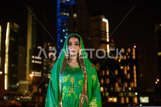 اظهار الولاء للبلاد، أزياء المناسبات والأعياد الوطنية، ذكرى اليوم الوطني السعودي 23 سبتمبر، يوم العلم 11 مارس، صورة مقربة لامرأة عربية خليجية سعودية مبتسمة ترتدي الشيلة وثوب المنطقة الوسطى في العيد الوطني، خلفية الابراج وناطحات السحاب المضيئة ليلا في الرياض