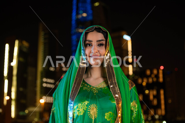 ايماءات العزه والفخر ،أزياء الاحتفالات الشعبيه بالأعياد والمناسبات الوطنية، ذكرى اليوم الوطني السعودي 23 سبتمبر، صورة مقربة لامرأة عربية خليجية سعودية مبتسمة ترتدي الشيلة وثوب المنطقة الوسطى في العيد الوطني، خلفية الابراج وناطحات السحاب المضيئة ليلا في الرياض