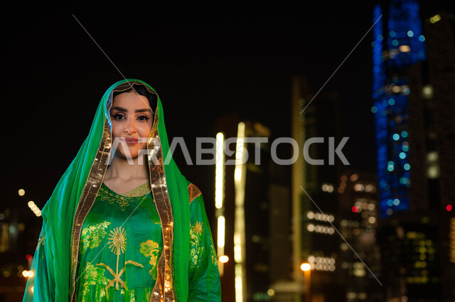 ايماءات الفخر والعزه ،أزياء الاحتفالات الشعبيه بالأعياد والمناسبات الوطنية، ذكرى اليوم الوطني السعودي 23 سبتمبر، صورة مقربة لامرأة عربية خليجية سعودية مبتسمة ترتدي الشيلة وثوب المنطقة الوسطى في العيد الوطني، خلفية الابراج وناطحات السحاب المضيئة ليلا في الرياض