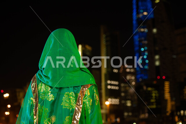 الولاء والاخلاص للسعودية ،أزياء الاحتفالات الشعبية في الأعياد والمناسبات الوطنية، ذكرى اليوم الوطني السعودي 8 ربيع الأول، صورة مقربة من الخلف لامرأة عربية خليجية سعودية ترتدي الشيلة وثوب المنطقة الوسطى في العيد الوطني، خلفية الابراج المضيئة ليلا في الرياض .