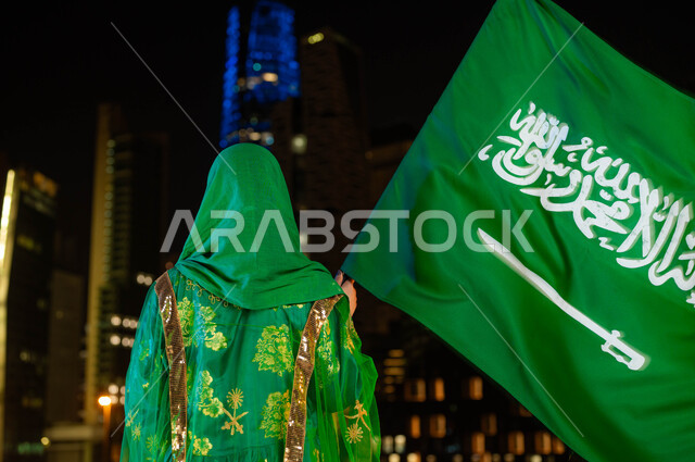 الوقوف أمام ناطحات السحاب المضيئة ليلا في الرياض، الاحتفال بذكرى اليوم الوطني السعودي 23 سبتمبر، مناسبات وطنية تقليدية، يوم العلم 11 مارس، صورة مقربة من الخلف لامرأة عربية خليجية سعودية ترتدي الشيلة وثوب المنطقة الوسطى في العيد الوطني تحمل بيدها علم المملكة