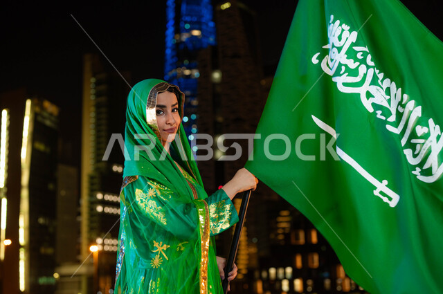 يوم العلم 11 مارس، الاحتفال بذكرى اليوم الوطني السعودي 23 سبتمبر، صورة مقربة من الجانب لامرأة عربية خليجية سعودية مبتسمة ترتدي الشيلة وثوب المنطقة الوسطى في العيد الوطني تحمل بيدها علم المملكة وتنظر إلى الكاميرا، خلفية ناطحات السحاب المضيئة ليلا في الرياض