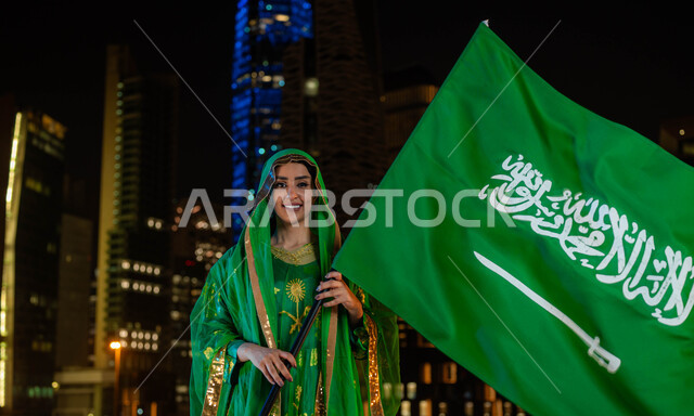 يوم العلم 11 مارس، ذكرى اليوم الوطني السعودي 23 سبتمبر، امرأة عربية خليجية مبتسمة سعودية ترتدي الشيلة وثوب المنطقة الوسطى في العيد الوطني  تحمل بيدها علم المملكة وتنظر إلى الكاميرا، احتفالات ومناسبات وطنية، خلفية الابراج وناطحات السحاب المضاءة ليلا في الرياض.