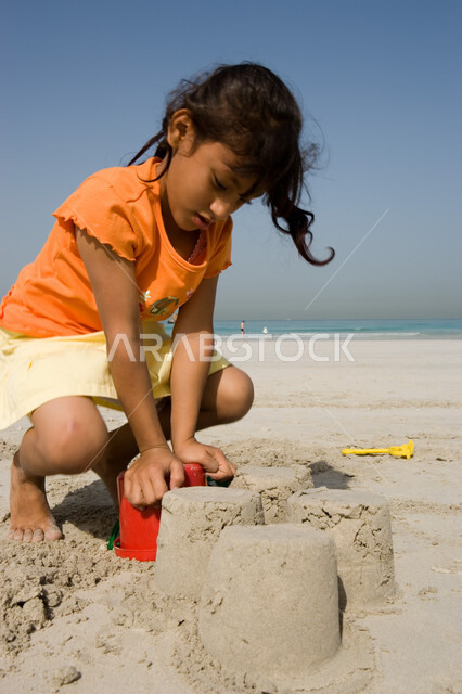 الاستمتاع باللعب بأدوات الرمل على ساحل البحر, قضاء أوقات صيفية ممتعة, صورة مقربة لفتاة صغيرة عربية خليجية اماراتية تقوم بصنع قلاع رملية على شاطئ البحر في إجازة الصيف، السياحة في دول الخليج العربي، نشاطات ترفيهية للأطفال
