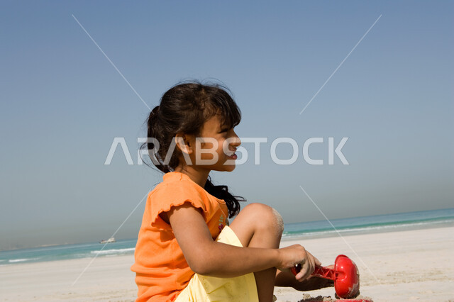 الاستمتاع باللعب بأدوات الرمل على ساحل البحر, قضاء أوقات صيفية ممتعة, صورة مقربة لفتاة صغيرة عربية خليجية اماراتية تقوم بصنع قلاع رملية على شاطئ البحر في إجازة الصيف، السياحة في دول الخليج العربي، نشاطات ترفيهية للأطفال