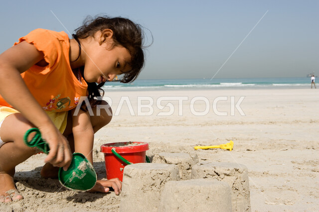قضاء أوقات صيفية ممتعة, الاستمتاع باللعب بأدوات الرمل على ساحل البحر, صورة مقربة لفتاة صغيرة عربية خليجية اماراتية تقوم بصنع قلاع رملية على شاطئ البحر في إجازة الصيف، السياحة في دول الخليج العربي، نشاطات ترفيهية للأطفال