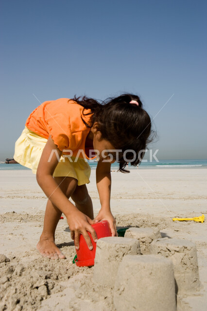 قضاء أوقات صيفية ممتعة, الاستمتاع باللعب بأدوات الرمل على ساحل البحر, صورة مقربة لفتاة صغيرة عربية خليجية اماراتية تقوم بصنع قلاع رملية على شاطئ البحر في إجازة الصيف، السياحة في دول الخليج العربي، نشاطات ترفيهية للأطفال