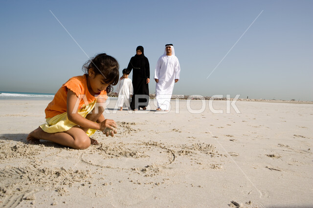 نشاطات ترفيهية للأطفال, قضاء أوقات صيفية ممتعة, الاستمتاع باللعب بالرمال على ساحل البحر مع العائلة, فتاة صغيرة عربية خليجية اماراتية تقوم بالرسم والكتابة على شاطئ البحر في إجازة الصيف، السياحة في دول الخليج العربي
