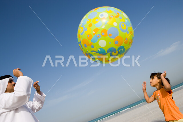 التسلية والترفيه مع الأبناء, قضاء أوقات صيفية ممتعة, الاستمتاع باللعب على ساحل البحر مع العائلة, أب عربي خليجي إماراتي يرتدي الغترة والكندورة الإماراتية يقوم باللعب مع ابنته بالكرة المطاطية على شاطئ البحر، السياحة في دول الخليج العربي