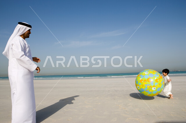 قضاء أوقات صيفية ممتعة بالتسلية و الترفيه مع الأبناء, الاستمتاع باللعب على ساحل البحر مع العائلة, أب عربي خليجي إماراتي يرتدي الغترة والكندورة الإماراتية يقوم باللعب مع ابنه بالكرة المطاطية على شاطئ البحر، السياحة في دول الخليج العربي