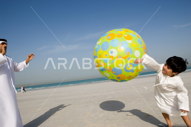 قضاء أوقات صيفية ممتعة بالتسلية و الترفيه مع الأبناء, الاستمتاع باللعب على ساحل البحر مع العائلة, أب عربي خليجي إماراتي يرتدي الغترة والكندورة الإماراتية يقوم باللعب مع ابنه بالكرة المطاطية على شاطئ البحر، السياحة في دول الخليج العربي