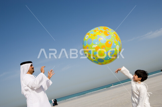 قضاء أوقات صيفية ممتعة بالتسلية و الترفيه مع الأبناء, الاستمتاع باللعب على ساحل البحر مع العائلة, أب عربي خليجي إماراتي يرتدي الغترة والكندورة الإماراتية يقوم باللعب مع ابنه بالكرة المطاطية على شاطئ البحر، السياحة في دول الخليج العربي