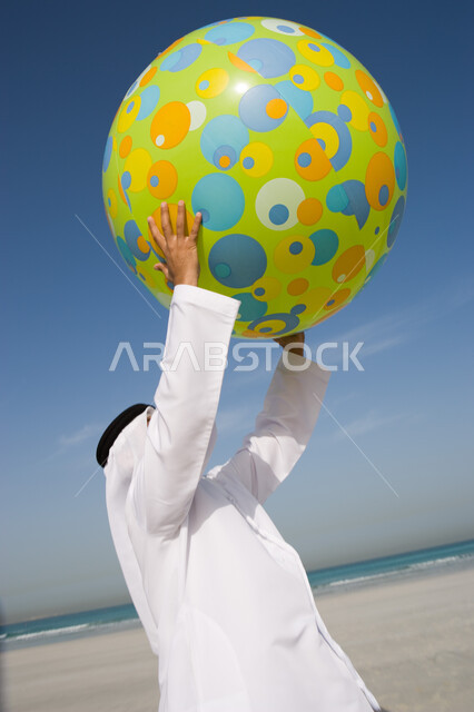 Beach resorts in the Emirates, spending pleasant summer times, enjoying playing on the sea coast during the summer vacation, an Emirati Arab man wearing a ghutra and an Emirati kandora playing with a rubber ball on the seashore, tourism in the Arab Gulf c
