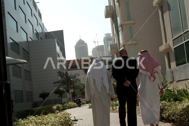انتهاء الدوام وخروج الموظفين, اجتماع عمل في الهواء الطلق, التجول ومناقشة الاعمال في الخارج, صورة من الخلف لرجلان عربيان خليجيان اماراتيان يرتديان الثوب الاماراتي ورجل بالبدلة الرسمية خارج مقر الشركه، شركات ومكاتب خليجيه في الإمارات العربيه المتحده