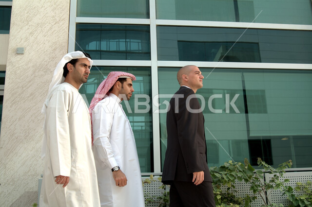 انتهاء الدوام وخروج الموظفين, اجتماع عمل في الهواء الطلق, التجول ومناقشة الاعمال في الخارج, صورة من الجانب لرجلان عربيان خليجيان اماراتيان يرتديان الثوب الاماراتي ورجل بالبدلة الرسمية خارج مقر الشركه، شركات ومكاتب خليجيه في الإمارات العربيه المتحده