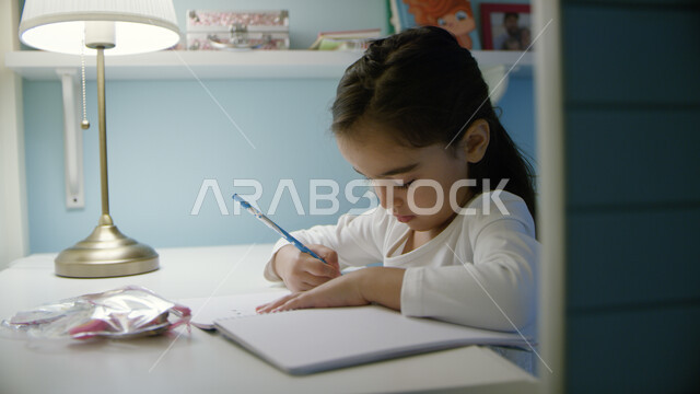 التعليم عن بعد، التحضير إلى الامتحان النصفي، الدراسة في الإمارات العربية المتحدة، صورة مقربة لفتاة صغيرة عربية خليجية إماراتية تجلس على الطاولة وتقوم بأداء الواجبات المدرسية، التعليم المنزلي أثناء الحجر الصحي و تفشي فايروس كورونا 