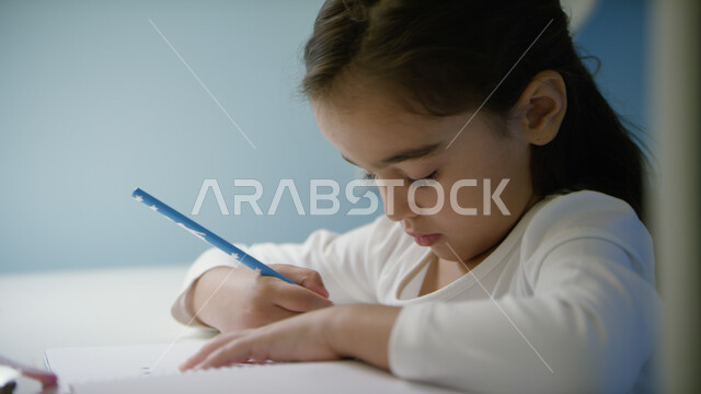 القيام بأداء الواجبات المدرسيه، التعليم عن بعد، الدراسة في الإمارات العربية المتحدة، صورة مقربة لفتاة صغيرة عربية خليجية إماراتية تجلس على الطالبة وتحمل بيدها قلم رصاص، التعليم المنزلي أثناء الحجر الصحي و تفشي فايروس كورونا