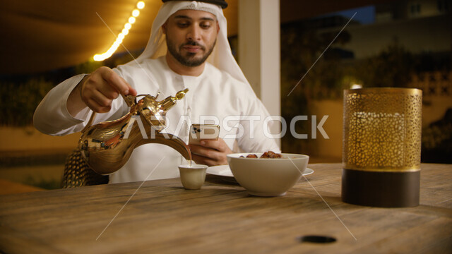 زينة و أضواء الشهر الفضيل, النظر إلى الوقت في الجوال و انتظار موعد الافطار, صورة مقربه لرجل عربي إماراتي خليجي يرتدي الغترة والكندورة الاماراتية يحمل بيده الهاتف المحمول ويقوم بصب القهوة ، تناول وجبة الفطور في إحدى المطاعم