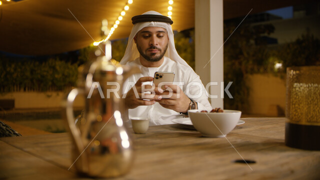 النظر إلى الوقت في الجوال و انتظار موعد الافطار, صورة مقربه لرجل عربي إماراتي خليجي يرتدي الغترة والكندورة الاماراتية يحمل بيده الهاتف المحمول ،تلقي الرسائل النصية وقت الإفطار، تناول وجبة الفطور في إحدى المطاعم, زينة و أضواء الشهر الفضيل