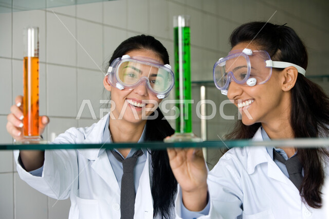 التعليم المتطور في جامعات الامارات العربية المتحدة, التطور والبحث والقيام باختبارات علمية, الجلوس في معمل الكيمياء, صورة مقربة لطالبتين عربيتين خليجيتين إماراتيتين مبتسمتين ترتديان المعطف الأبيض ونظارات الحماية تقومان بتجربة كيميائية