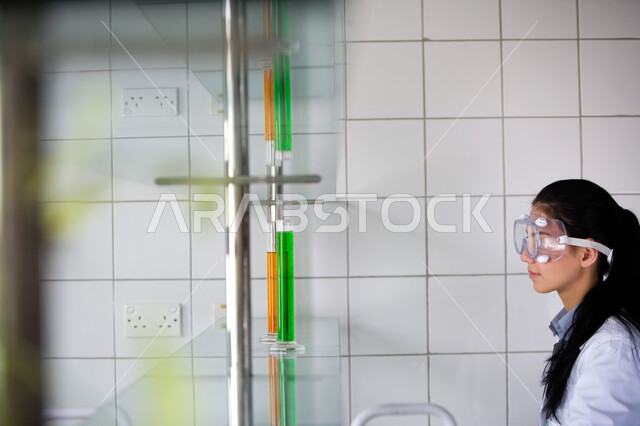 البحث والتطور والقيام باختبارات علمية, إجراء التجارب الكيميائية في المختبر باستخدام أنابيب الاختبار, الجلوس في معمل الكيمياء, صورة من الجانب لطالبة جامعية عربية خليجية إماراتية ترتدي المعطف الأبيض ونظارات الحماية تمسك بيدها الأنبوب الزجاجي