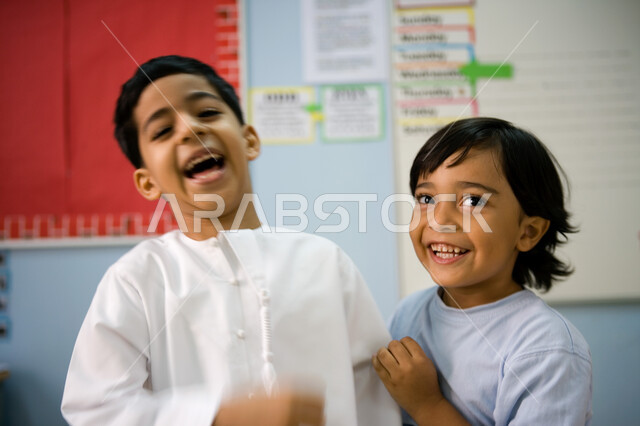 An enthusiastic and happy school atmosphere, laughter and smiles in the classroom, a close-up of two Arab Gulf Emirati students inside the classroom at school, merging with friends, enjoying spending quality time with gestures of pleasure, happiness and j