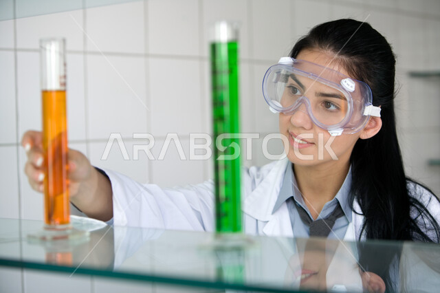 Sitting in the chemistry laboratory, developing, researching and doing scientific tests, conducting chemical experiments in the laboratory using test tubes, a close-up of an Arab Gulf Emirati university student wearing a white coat and protective glasses 