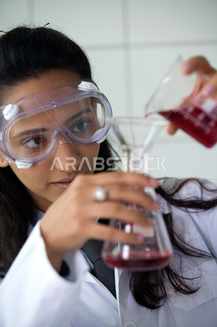 إجراء التجارب الكيميائية في المختبر باستخدام أنابيب الاختبار, الجلوس في معمل الكيمياء, صورة مقربة لطالبة جامعية عربية خليجية إماراتية ترتدي المعطف الأبيض ونظارات الحماية تمسك بيدها الأنبوب الزجاجي, التطور والبحث والقيام باختبارات علمية