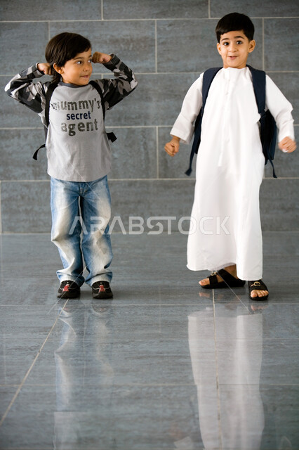 اللعب في بهو المدرسة, تلميذان عربيان خليجيان إماراتيان يرتديان حقيبة الظهر المدرسية, ايماءات القوة والحماس بالعودة إلى المدرسة, الاستمتاع بقضاء وقت ممتع مع الأصدقاء, المدارس الأكاديمية في الإمارات العربية المتحدة, صورة الجسم بالطول الكامل