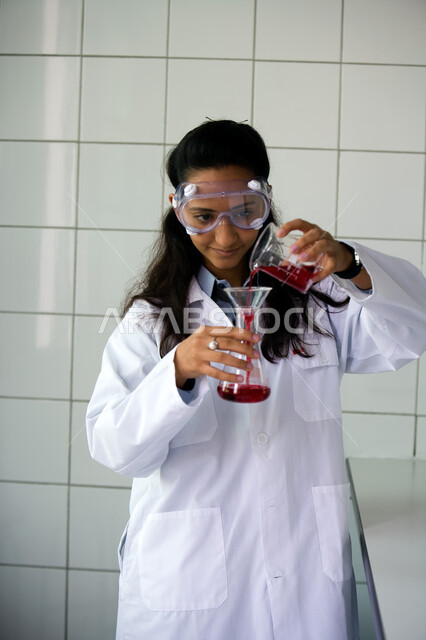 التطور والبحث والقيام باختبارات علمية, إجراء التجارب الكيميائية في المختبر باستخدام أنابيب الاختبار, الجلوس في معمل الكيمياء, صورة مقربة لطالبة جامعية عربية خليجية إماراتية ترتدي المعطف الأبيض ونظارات الحماية تمسك بيدها الأنبوب الزجاجي