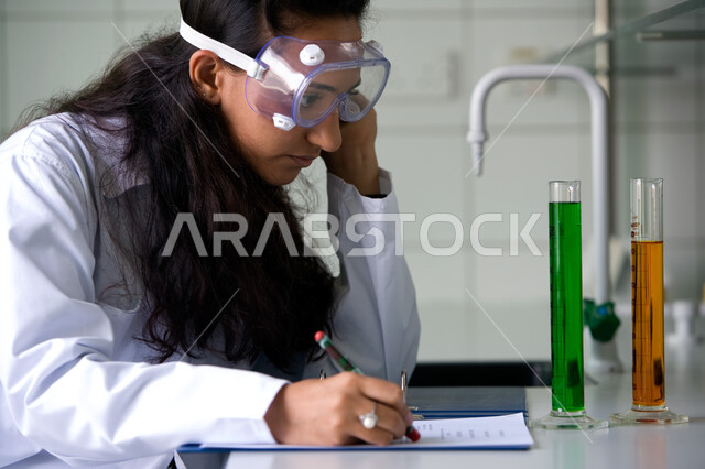 النظر إلى شيء ما بايماءات التفكير والتركيز,  الجلوس في مختبرات ومعامل الكيمياء, صورة مقربة لطالبة عربية خليجية إماراتية ترتدي المعطف الأبيض ونظارة الوقاية تقوم بكتابة تقرير عن المحاليل والسوائل والاختبارات الكيميائية