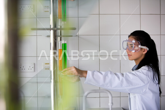 التطور والبحث والقيام باختبارات علمية, إجراء التجارب الكيميائية في المختبر باستخدام أنابيب الاختبار, الجلوس في معمل الكيمياء, صورة من الجانب لطالبة جامعية عربية خليجية إماراتية ترتدي المعطف الأبيض ونظارات الحماية تمسك بيدها الأنبوب الزجاجي