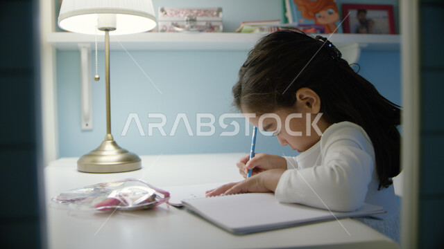 التحضير إلى الامتحان النصفي، التعليم عن بعد، الدراسة في الإمارات العربية المتحدة، صورة مقربة لفتاة صغيرة عربية خليجية إماراتية تجلس على الطالبة وتقوم بأداء الواجبات المدرسية، التعليم المنزلي أثناء الحجر الصحي و تفشي فايروس كورونا 
