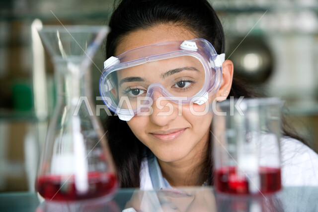 تطور طرق التعليم والقيام بتجارب كيميائية متطورة بإيماءات الدقة والتركيز، صورة مقربة لطالبة عربية خليجية اماراتية مبتسمة ترتدي نظارة الوقاية والمعطف الأبيض تنظر إلى الأنابيب الزجاجية، اكتساب المعرفة والاستفادة من الأبحاث المخبرية الحديثة