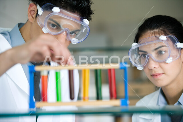 المشاركة بين الطلاب والاستفادة من الأبحاث المخبرية, استخدام أنابيب الاختبار لإجراء التجارب في المختبر, صورة مقربة لشاب وفتاة عربيين خليجيين إماراتيين ينظران لأنبوب الاختبار بإيماءات التركيز والدقة, ارتداء النظارة الوقائية والقيام بتجربة كيميائية