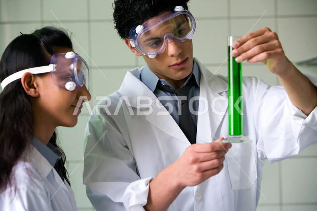 الاستفادة من الأبحاث المخبرية والمشاركة بين الزملاء , استخدام أنابيب الاختبار لإجراء التجارب في المختبر, صورة مقربة لشاب وفتاة عربيين خليجيين إماراتيين في بيئة علمية متطورة, ارتداء النظارة الوقائية والقيام بتجربة كيميائية بإيماءات التركيز والدقة
