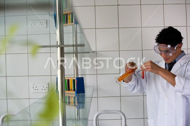 التطور والبحث وإجراء التفاعلات الكيميائية, طالب جامعي عربي خليجي إماراتي يقوم بتجربة علمية في المختبر, ارتداء نظارة الوقاية والمعطف الأبيض, اكتساب المعرفة والخبرة بإيماءات الدقة والتركيز في التجربة, الدراسات والتجارب في بيئة عملية متطورة