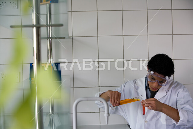 البحث والتطوير وإجراء التفاعلات الكيميائية, طالب جامعي عربي خليجي إماراتي يقوم بتجربة علمية في المختبر, ارتداء نظارة الوقاية والمعطف الأبيض, اكتساب المعرفة والخبرة بإيماءات الدقة والتركيز في التجربة, الدراسات والتجارب في بيئة عملية متطورة