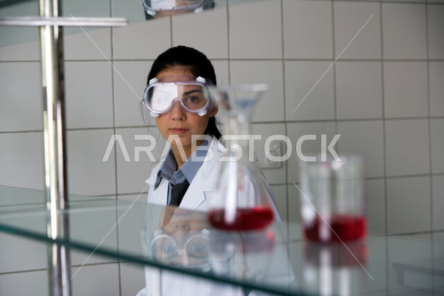الحرص في إجراء التجارب العلمية, طالبة عربية خليجية إماراتية ترتدي نظارة الوقاية والمعطف الأبيض تنظر إلى الأنابيب الزجاجية, التفاعلات الكيميائية داخل المختبر, انتظار نتيجة الدراسات وتحاليل المواد الكيميائية, مفهوم الكيمياء الحيوية