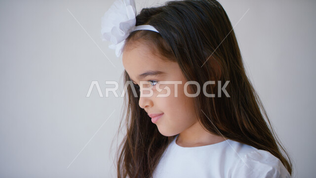 Looking at something with thinking gestures and absent-mindedness, taking care of the external appearance of young children, a close-up side portrait of a smiling Emirati Gulf Arab girl wearing new clothes on holidays and occasions, a white background