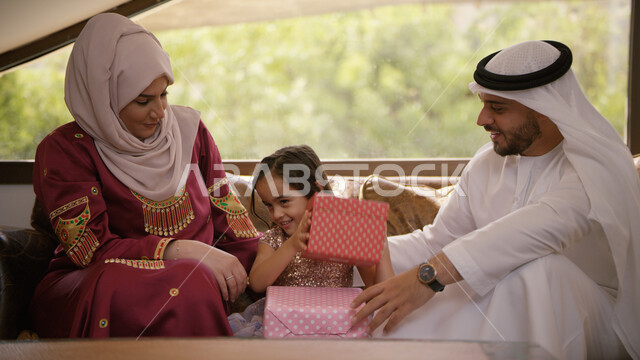 ايماءات التفكير والتخمين في محتوى الهديه, أجواء عائلية سعيدة مليئة بالحب, الاحتفال بقدوم العيد المبارك، عائلة عربية خليجية اماراتيه يجلسون في غرفة المعيشة بإيماءات السعادة والفرح، ارتداء الزي الإماراتي وتقديم الهدايا في الأعياد والمناسبات الإسلامية