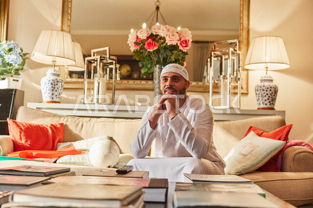 ايماءات الانتظار والتشوق لتسجيل الهدف، رجل عربي خليجي اماراتي يرتدي الكندورة والطاقية البيضاء الاماراتية يجلس على الاريكة يقوم بمتابعة مباراة كرة القدم، تشجيع المنتخب الوطني، غرفة معيشة مليئة بالكتب التعليمية الثقافية