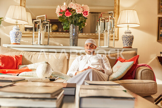 The impact of the failure and loss of the favorite team on the fan, watching the football match, an Arab Emirati man wearing a kandora and the Emirati white cap sitting on the sofa with gestures of sadness and misery, encouraging the national team, a livi